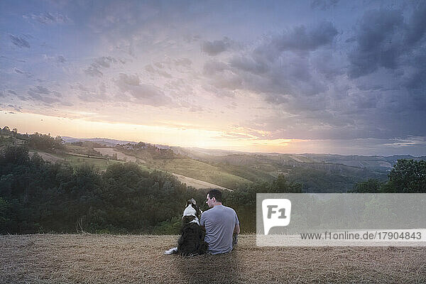 Ein Mann und sein Border Collie Hund sitzen auf einem Feld in der Landschaft und genießen den Sonnenuntergang  Italien  Europa