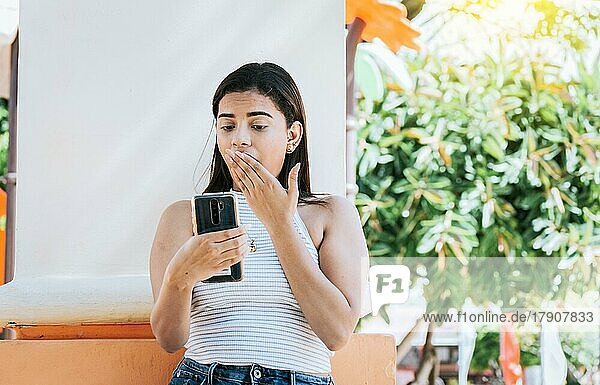 Mädchen mit Handy mit einem überraschten Gesicht lehnt an einer Wand. Junge Frau mit dem Handy mit einer Geste der Überraschung bedeckt ihren Mund im Freien. Überrascht Person Blick auf Handy