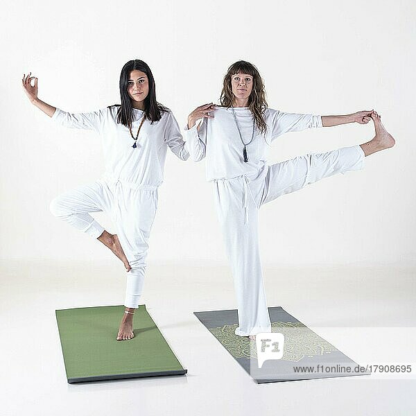 Zwei weiß gekleidete Frauen machen Yoga-Posen  während sie in die Kamera auf weißem Hintergrund schauen. Studioaufnahme