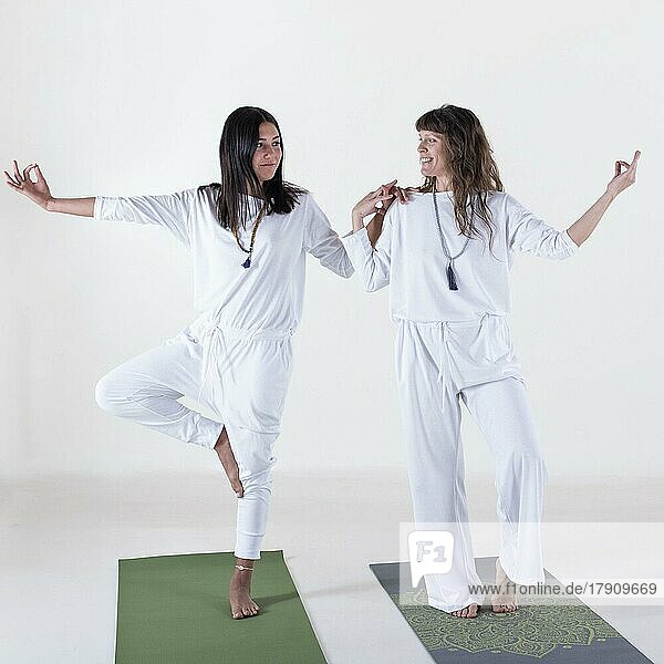 Zwei weiß gekleidete Frauen machen Yoga vor weißem Hintergrund. Die eine unterrichtet die andere. Studioaufnahme