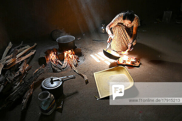 Sonnenlicht fällt in eine traditionelle Küche mit Lehmwänden und Holzfeuer  eine Frau macht Chai  Reissieb aus Plastik  Kadana  Gujarat  Indien  Asien