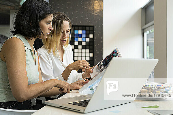 Side view shot of two female interior designers at their desk working with books and a laptop computer
