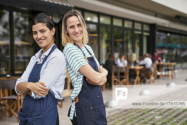 Foto von zwei glücklichen Restaurantbesitzerinnen  die Rücken an Rücken vor ihrem Restaurant stehen