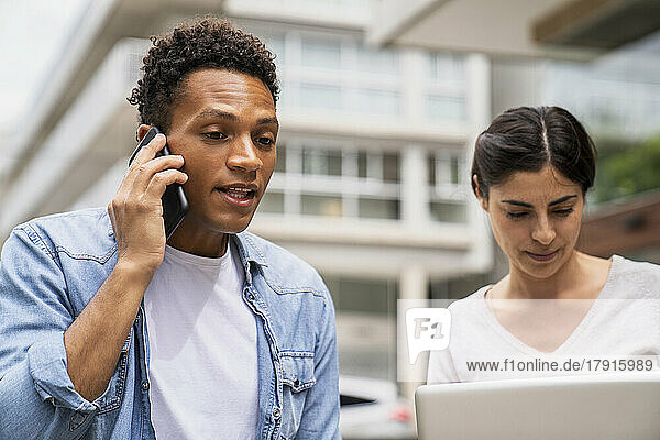 Mittelaufnahme eines afroamerikanischen Mannes  der mit einem Mobiltelefon spricht  und einer lateinamerikanischen Frau  die im Freien an einem Laptop-Computer arbeitet
