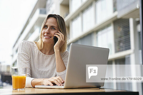 Porträt einer amüsierten Frau  die im Freien mit einem Laptop sitzt  während sie mit einem Handy telefoniert und Orangensaft trinkt
