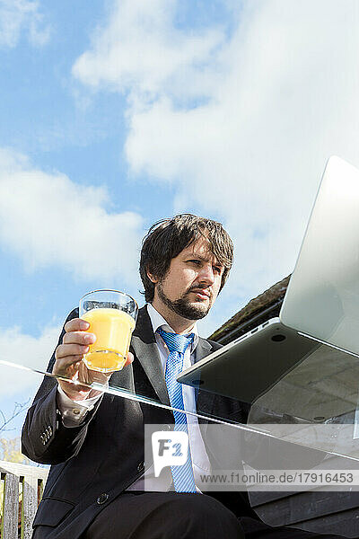 Ein Mann hält ein Glas Saft in der Hand und arbeitet draußen an einem Glastisch.