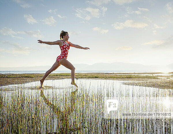 Girl (12-13) in swimsuit jumping above lake at sunrise