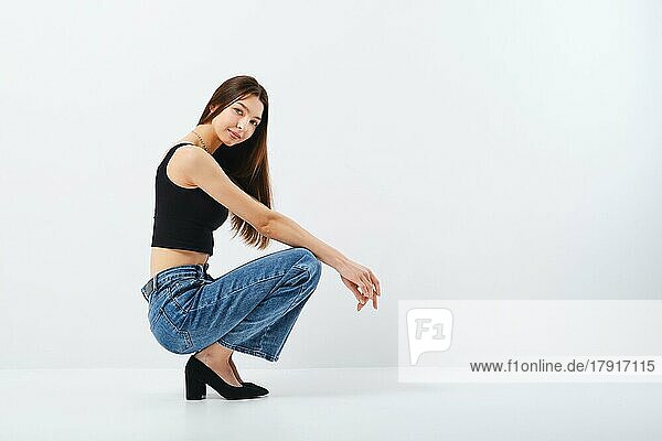 Ganzkörperporträt einer jungen Frau in schwarzem Tanktop und weiten Jeans im Profil sitzend