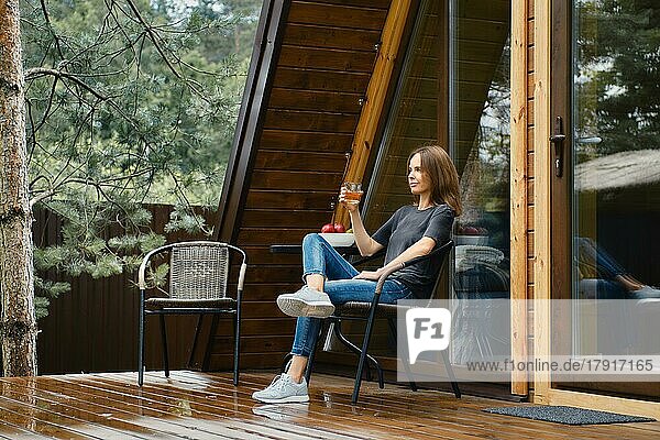 Süße Frau sitzt auf der Terrasse ihres Waldhauses und trinkt Saft