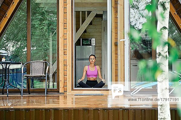 Frau sitzt im Yoga-Lotus-Sitz im Waldhaus vor geöffneter Tür und genießt die frische Luft nach dem Regen