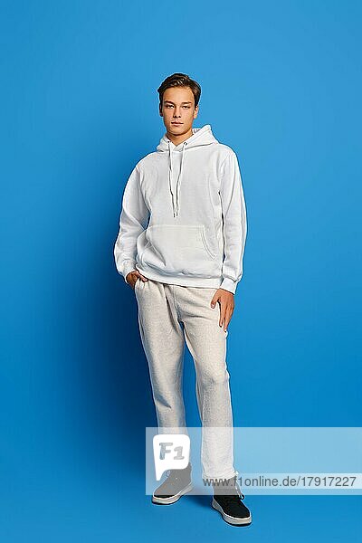 Attraktiver stilvoller Mann in weißem Sweatshirt und Jogginghose vor blauem Studiohintergrund