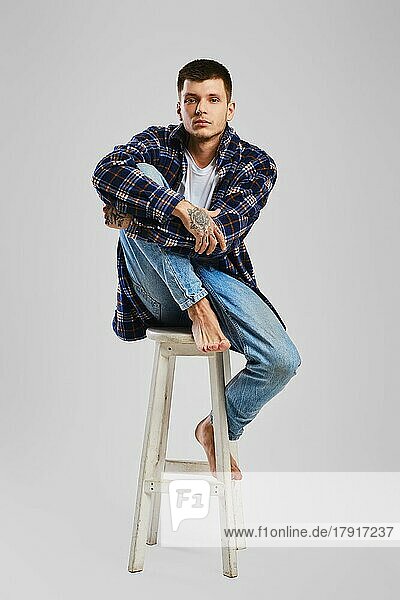 Ganzes Studioporträt eines jungen barfüßigen Mannes in Hemd und Jeans  der auf einem hohen Holzstuhl sitzt