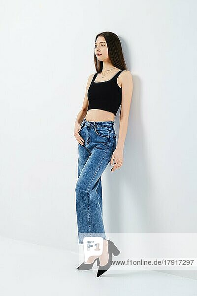 Ganzkörperporträt einer jungen Frau in schwarzem Tank-Top und Denim-Jeans  die sich an die weiße Studiowand lehnt  Seitenansicht