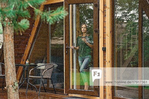 Junge Frau mit einer Tasse Kräutertee steht auf der Türschwelle einer Waldhütte und schaut auf den Regen draußen