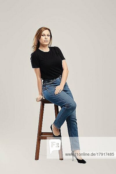 Junge Frau in schwarzem T-Shirt und Jeans posiert auf hohem Holzstuhl vor grauem Hintergrund