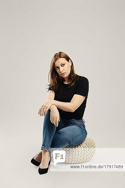 Nachdenkliche Frau in lässiger schwarzer Bluse und Jeans sitzt auf dem Studioboden vor grauem Hintergrund