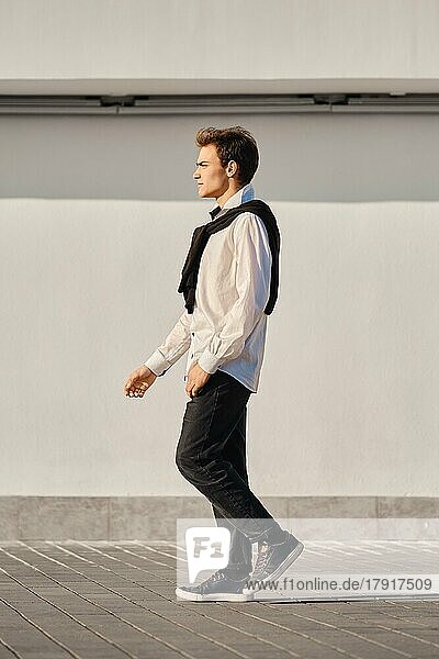 Ganzkörperporträt eines jungen Mannes in weißem Hemd und über die Schultern gezogenem Pullover geht auf der Straße