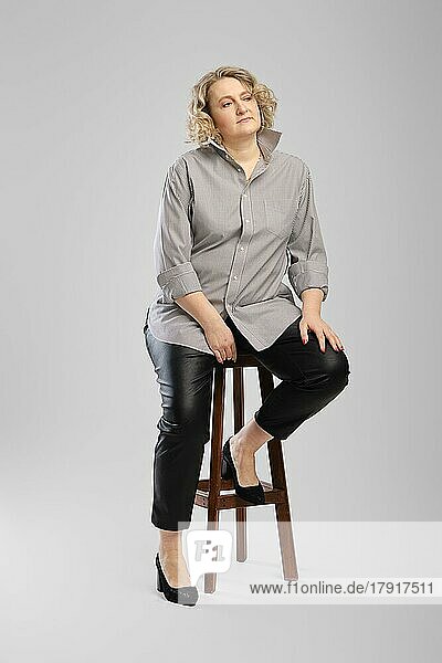 Ältere Frau in grauem Hemd und Kunstlederhose sitzt auf einem hohen Holzstuhl vor grauem Studiohintergrund