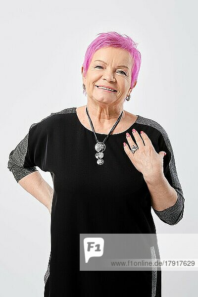 Positive alte Frau mit kurzen rosa Haaren erklärt und gestikuliert etwas in weißem Studio