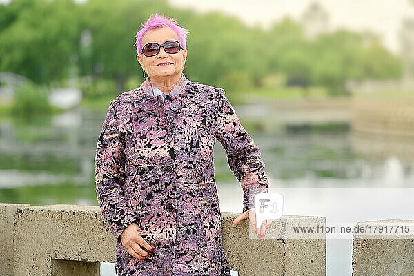 Aktive ältere Frau mit fröhlichem Lächeln steht am Flussufer im Stadtpark