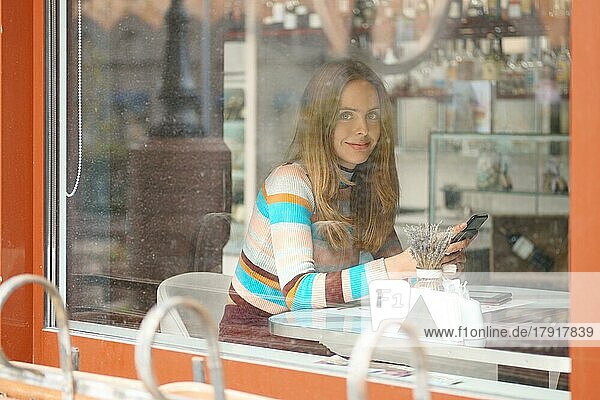 Nettes Mädchen mit langen Haaren sitzt im Café und plaudert mit dem Handy. Foto von außen aufgenommen  mit einem Blick durch das Fenster
