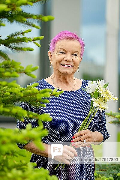 Porträt einer lächelnden reifen Frau mit kurzen rosafarbenen Haaren  die in der Nähe eines Tannenbaums mit Frühlingsblumen in den Händen im Freien steht