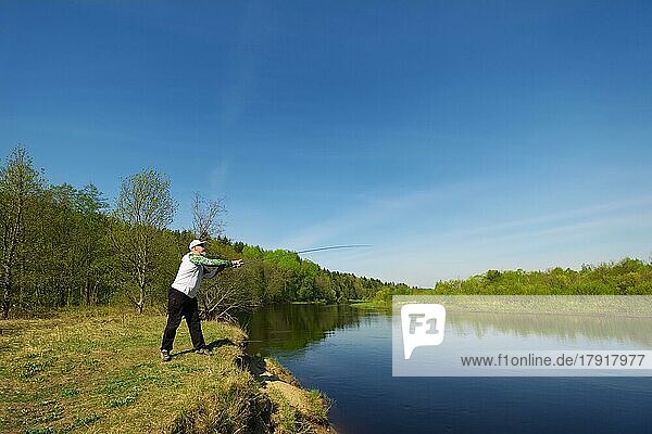 Angler mit einer Spinnrute fangen Fische auf einem Fluss an einem sonnigen Sommertag mit grünen Bäumen im Hintergrund. Wochenendaktivität im Freien. Foto mit geringer Schärfentiefe bei weit geöffneter Blende aufgenommen