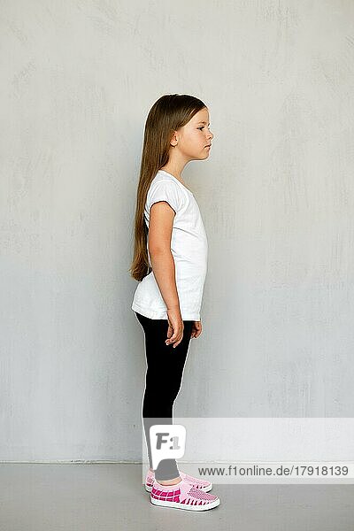 Niedliches kleines Kind mit langen Haaren in weißem T-Shirt und schwarzer Jogginghose im Profil stehend