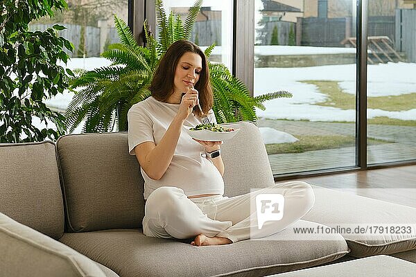Schwangere Frau  die sich um ihr Kind und ihre Gesundheit kümmert  sitzt auf dem Sofa im Wohnzimmer und isst frischen Gemüsesalat