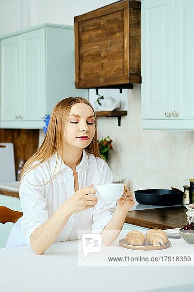 Nachdenkliche junge Frau sitzt hinter dem Küchentisch mit einer Tasse Kaffee und denkt über etwas nach
