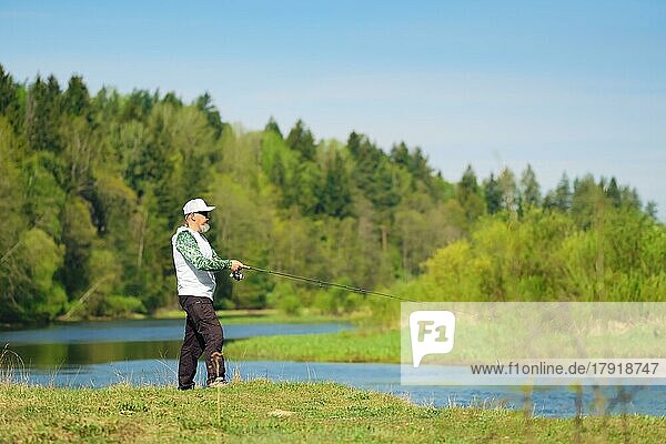 Fisherman with a spinning rod catching fish on a river at sunny summer day with green trees at background. Outdoor weekend activity. Photo with shallow depth of field taken at wide open aperture