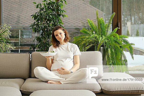 Schöne schwangere Frau sitzt auf dem Sofa im Wohnzimmer und hält im Arm frischen Gemüsesalat