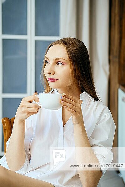 Nachdenkliche junge Frau im weißen Hemd in der Küche hält eine Tasse Kaffee
