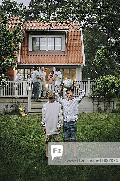 Playful Jungen mit Fußball stehen in Hinterhof mit Familie auf Veranda im Hintergrund