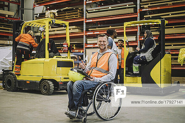 Full length portrait of smiling carpenter with digital tablet sitting on wheelchair in warehouse
