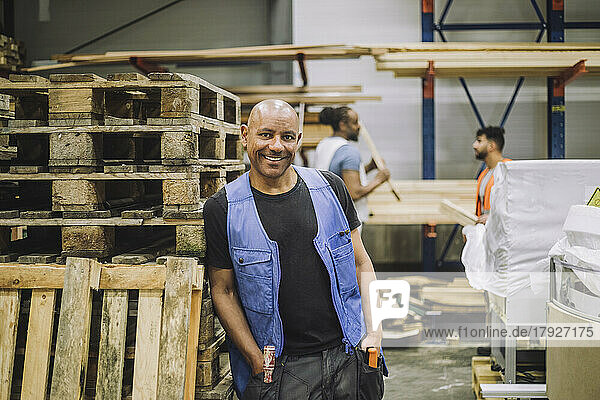 Smiling bald carpenter with hands in pockets leaning on wooden rack in warehouse
