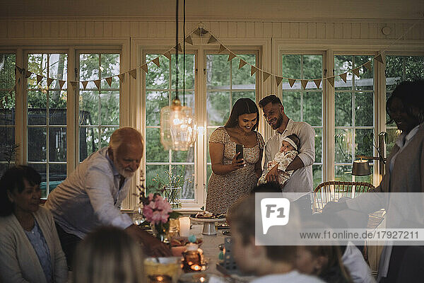 Glückliche Mutter zeigt ihrer Tochter  die vom Vater gehalten wird  das Smartphone während einer Dinnerparty zu Hause