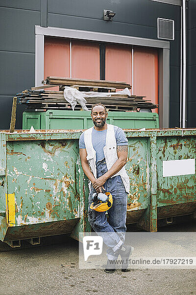 Lächelnder männlicher Bauarbeiter mit Schutzhelm in voller Länge  der sich auf der Baustelle an einen Metallcontainer lehnt
