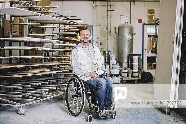 Lächelnder Maler in voller Länge  der einen Gehörschutz trägt  während er auf einem Rollstuhl in einer Werkstatt sitzt