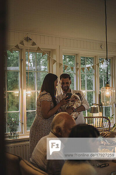 Frau  die ihr Smartphone mit einem lächelnden Mann teilt  der seine Tochter während einer Dinnerparty zu Hause hält
