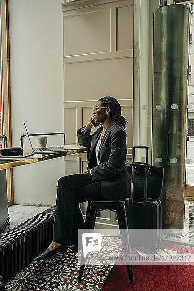 Full length of smiling female entrepreneur talking on mobile phone while sitting with laptop in hotel lounge