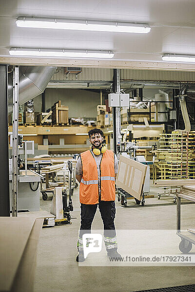 Full length of smiling carpenter wearing reflective clothing and ear protectors in warehouse