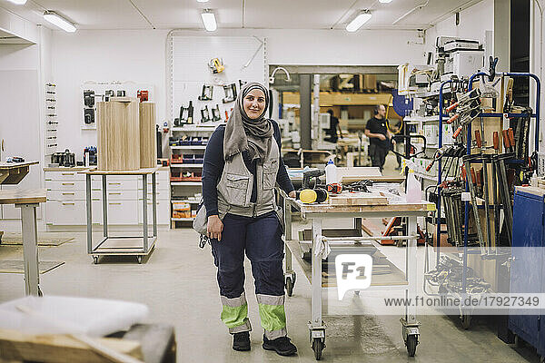 Full length portrait of smiling female carpenter standing by workbench in workshop