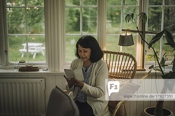 Reife Frau  die ihr Smartphone benutzt  während sie auf einem Stuhl in der Nähe des Fensters zu Hause sitzt