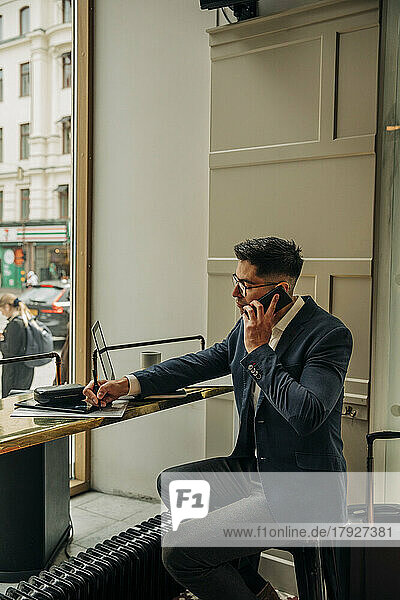 Geschäftsmann  der am Tisch in der Hotellounge sitzt und schreibt  während er mit seinem Smartphone telefoniert