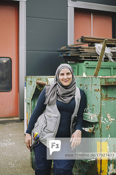 Lächelnde Bauarbeiterin mit Kopftuch lehnt sich an einen Metallcontainer