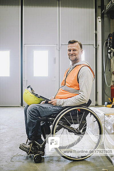 Seitenansicht eines Zimmermanns mit Schutzhelm und digitalem Tablet im Rollstuhl sitzend im Lager