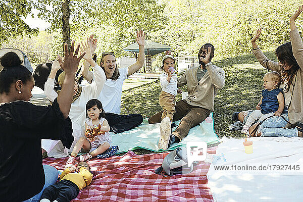 Glückliche Eltern mit erhobenen Armen sitzen mit Kindern auf einer Picknickdecke im Park