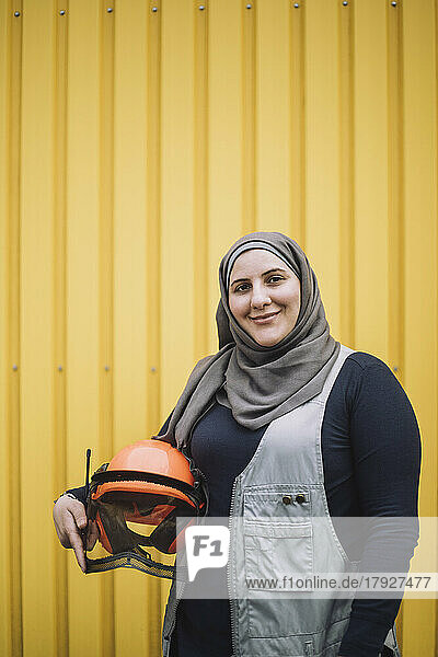 Porträt einer lächelnden Bauarbeiterin mit Kopftuch und Schutzhelm an einer gelben Metallwand