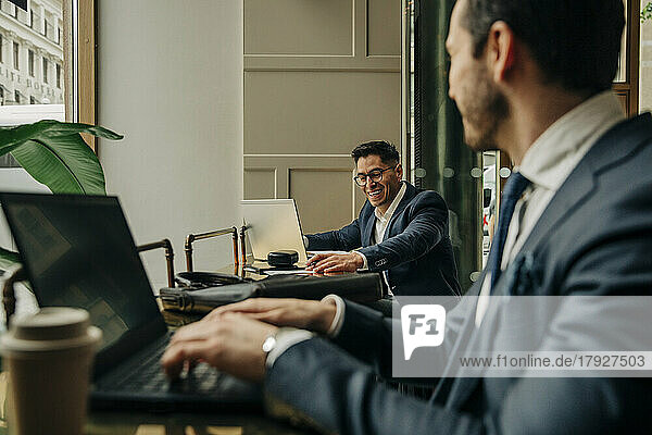 Happy businessman talking with male colleague using laptop while sitting in hotel lounge